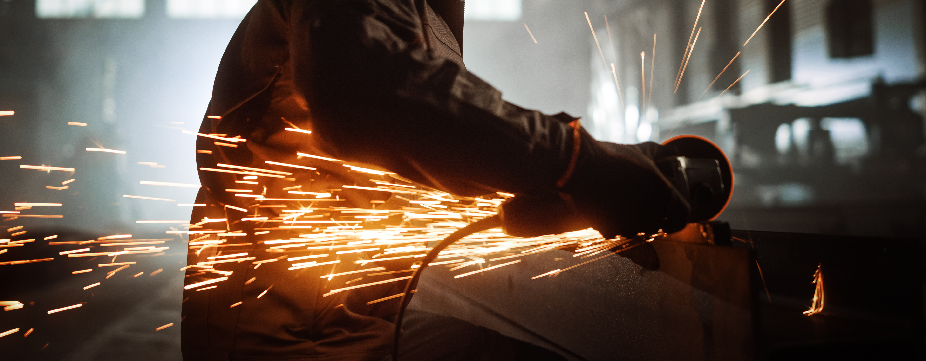 Heavy Industry Engineering Factory Interior with Industrial Worker Using Angle Grinder and Cutting a Metal Tube. Contractor in Safety Uniform and Hard Hat Manufacturing Metal Structures.